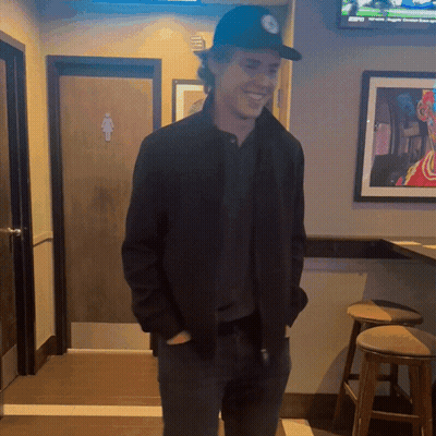 Man In Black Hat Smiling In Front Of Bathroom Door GIF