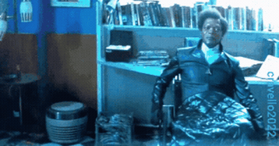 Man In Black Jacket Sitting By Bookshelf GIF