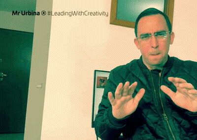 Man In Black Jacket Sitting In Front Of Mr Urbina #LeadingWithCreativity Sign GIF
