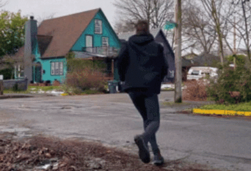 Man In Black Jacket Walking Past Blue House GIF