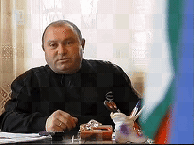 Man In Black Shirt At Desk With Pen Holder In Front Of Flag GIF
