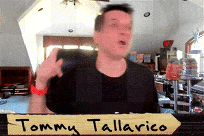 Man In Black Shirt In Front Of Tommy Tallarico Sign GIF