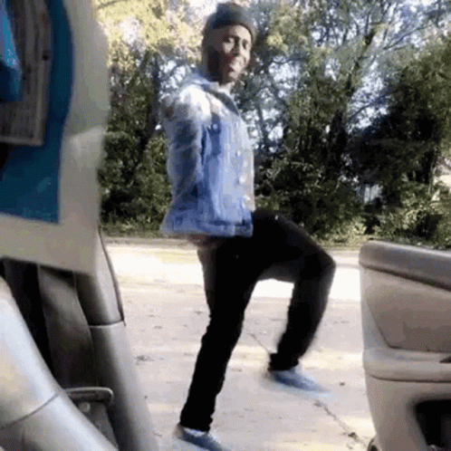 Man In Blue Denim Jacket Standing On One Leg In Front Of Car GIF