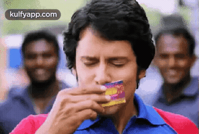 Man In Blue Shirt Eating Candy Bar While Men Laugh GIF