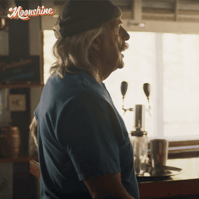 Man In Blue Shirt In Front Of Moonshine Sign GIF
