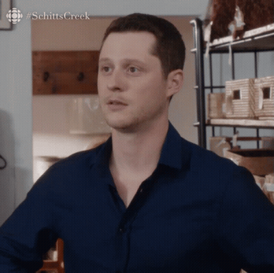 Man In Blue Shirt In Front Of Schitts Creek Shelf GIF