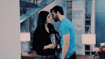 Man In Blue Shirt Kissing Woman On Cheek In Front Of Staircase GIF