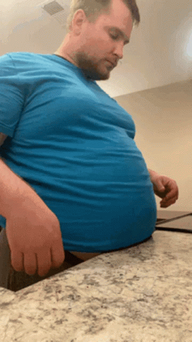 Man In Blue Shirt Sitting On Counter With Hands On Stomach GIF