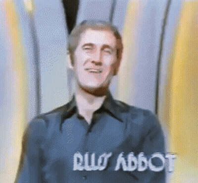 Man In Blue Shirt Smiling In Front Of Rus Abbot Sign GIF