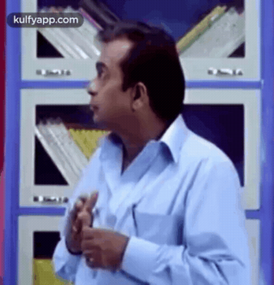 Man In Blue Shirt Standing In Front Of Bookshelf With Hands In Pockets GIF