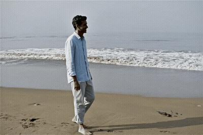 Man In Blue Shirt Walking Barefoot On Beach GIF
