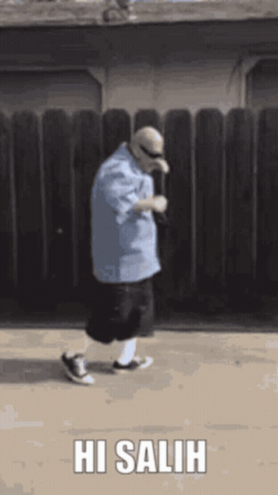 Man In Blue Shirt Walking In Front Of Wooden Fence Saying Hi Salih GIF