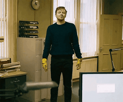 Man In Blue Sweater And Yellow Gloves Standing Before Clock GIF