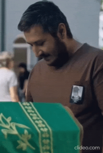 Man In Brown Shirt Looking At Green Coffin With Portrait GIF