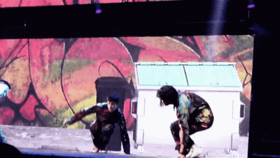 Man In Colorful Outfit Squatting By Dumpster GIF
