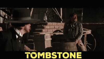 Man In Cowboy Hat Holding Gun In Front Of Tombstone GIF
