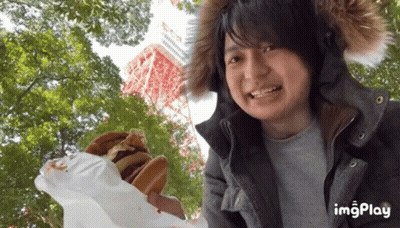 Man In Fur Hood Holding Hamburger In Front Of Tower And Trees GIF