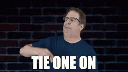 Man In Glasses And Blue Shirt Says Tie One On In Front Of Brick Wall GIF