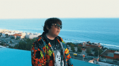 Man In Glasses And Colorful Jacket On Balcony Overlooking City And Ocean GIF