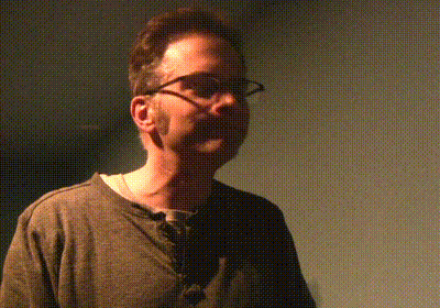 Man In Glasses And Grey Shirt Standing In Dark Room GIF