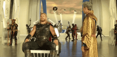 Man In Gold Robe Standing Next To Bearded Man In Chair GIF