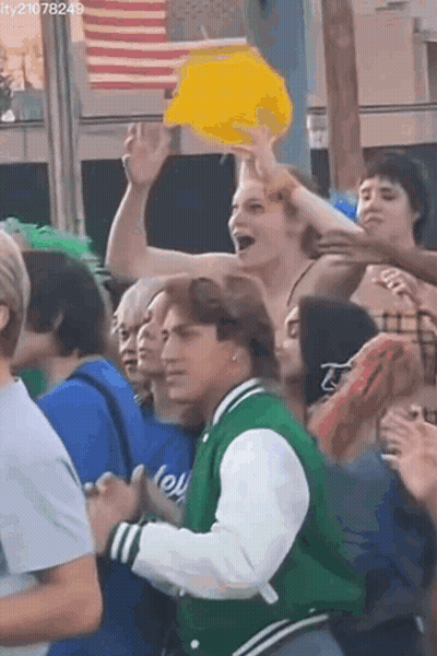 Man In Green Jacket Holding Yellow Hat In Crowd GIF