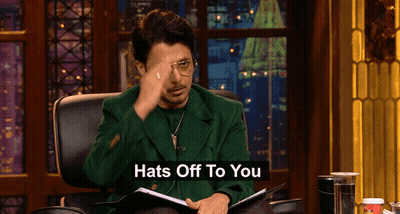 Man In Green Jacket Sitting With Hats Off Sign GIF