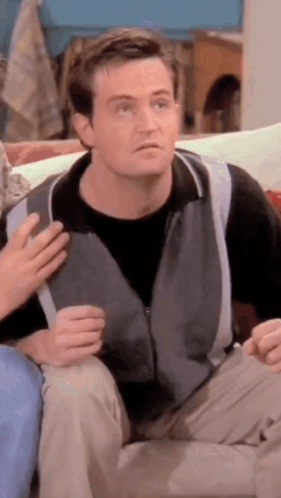 Man In Grey Vest Sitting On Couch Talking To Another Man GIF
