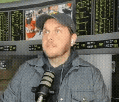 Man In Hat And Jacket Talking Into Microphone At Betting Board GIF