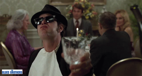 Man In Hat And Sunglasses Holding Glass In Front Of Blues Brothers Poster GIF