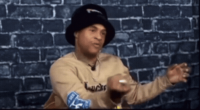 Man In Hat And Sweater Sitting By Brick Wall Talking GIF