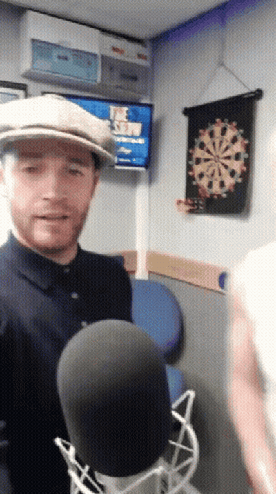 Man In Hat At Microphone In Front Of Dart Board GIF