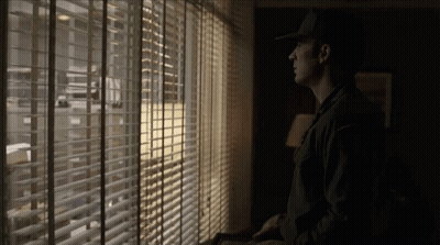 Man In Hat Looking Out Window With Blinds GIF