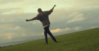 Man In Hat On Grassy Hill With Arms Outstretched GIF