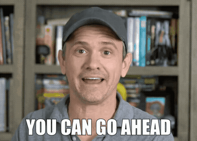 Man In Hat Saying You Can Go Ahead In Front Of Bookshelf GIF