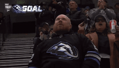 Man In Hockey Jersey Laying On Bleachers Watching Game With Scoreboard Showing Goal GIF