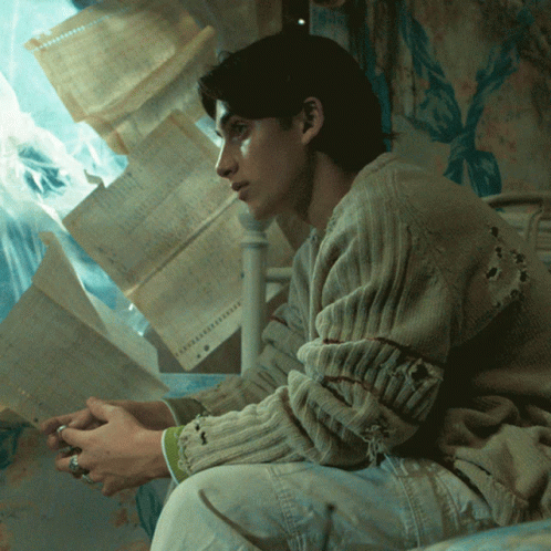 Man In Hole Sweater Sitting On Bed With Papers GIF