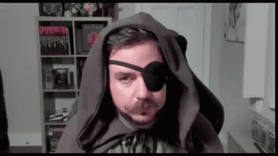 Man In Hooded Cape And Eye Patch Sitting In Front Of Bookshelf GIF