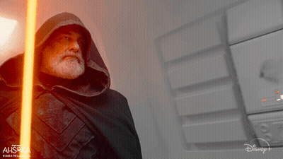 Man In Hooded Cape Holding Lightsaber In Front Of Ahsoka Sign GIF