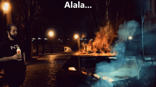 Man in N7 Shirt in Front of Burning Car at Night