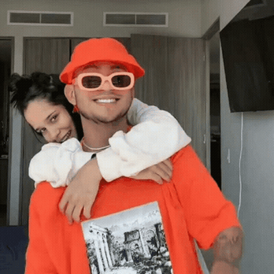 Man In Orange Hat And Sunglasses Carrying Woman On Back GIF