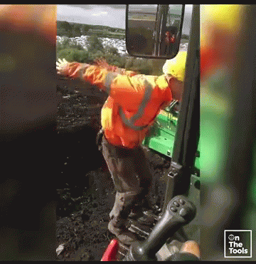 Man In Orange Jacket And Yellow Hard Hat In Front Of Green Tractor With Arms Outstretched GIF