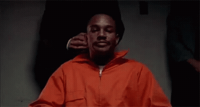 Man In Orange Prison Uniform Sitting With Man Holding His Ear GIF