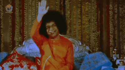 Man In Orange Shirt Sitting On Bed Waving GIF
