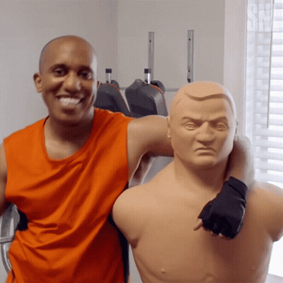 Man In Orange Tank Top Posing With Boxing Mannequin GIF