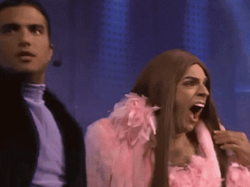 Man In Pink Feathered Coat Next To Woman In Wig GIF