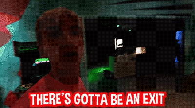 Man In Pink Shirt Standing In Dark Room With Sign Somewhere Over Here GIF