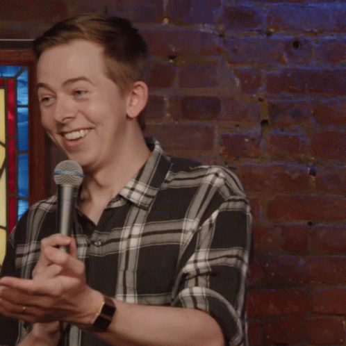 Man In Plaid Shirt Holding Microphone In Front Of Stained Glass Window GIF