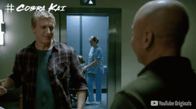 Man In Plaid Shirt Talking To Another Man In Front Of Elevator With Cobra Kai Written On Wall GIF