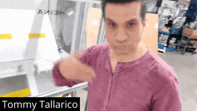 Man In Purple Shirt Standing Before Tommy Tallarico Sign GIF